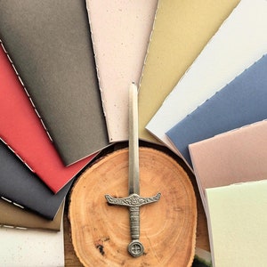 May include: A selection of small, colourful notebooks with stitched spines, arranged around a decorative sword on a wooden slice. The notebooks are in various colours, including red, grey, and blue. The sword is silver-toned.