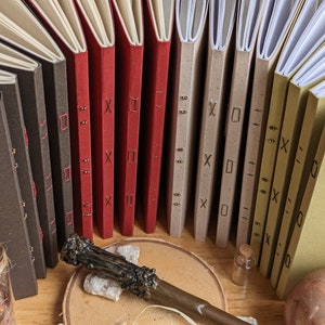May include: A collection of handmade journals with different coloured covers, including brown, red, beige, and green. The journals are bound with decorative stitching and have unique markings on the covers.