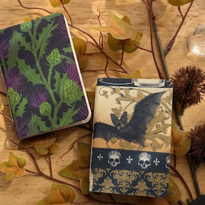 May include: Two small notebooks. One features a dark background with green leaves and purple thistle flowers. The other has a bat illustration with a decorative border of skulls and fleur-de-lis. Both are on a wooden surface.
