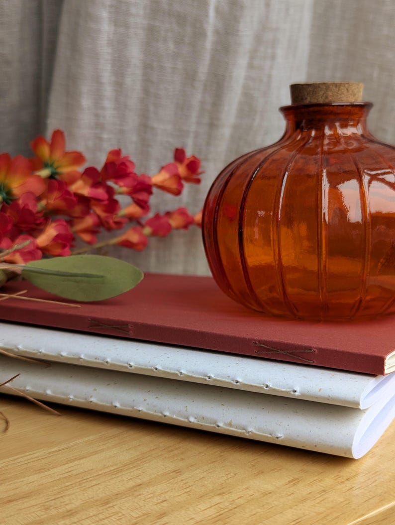May include: A stack of two hand-bound notebooks with visible stitching along the spines, one in terracotta red and one in speckled white, topped with a ribbed amber glass bottle featuring a cork stopper and a sprig of orange and red flowers.