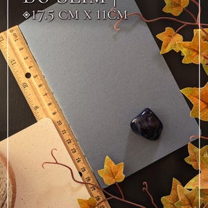 May include: A rectangular grey B6 Slim notebook measuring 17.5 cm by 11 cm, positioned next to a wooden ruler and a small, polished dark blue stone. The notebook is displayed with a wooden ruler showing metric measurements along its side.