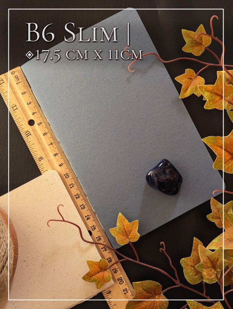 May include: A rectangular grey B6 Slim notebook measuring 17.5 cm by 11 cm, positioned next to a wooden ruler and a small, polished dark blue stone. The notebook is displayed with a wooden ruler showing metric measurements along its side.