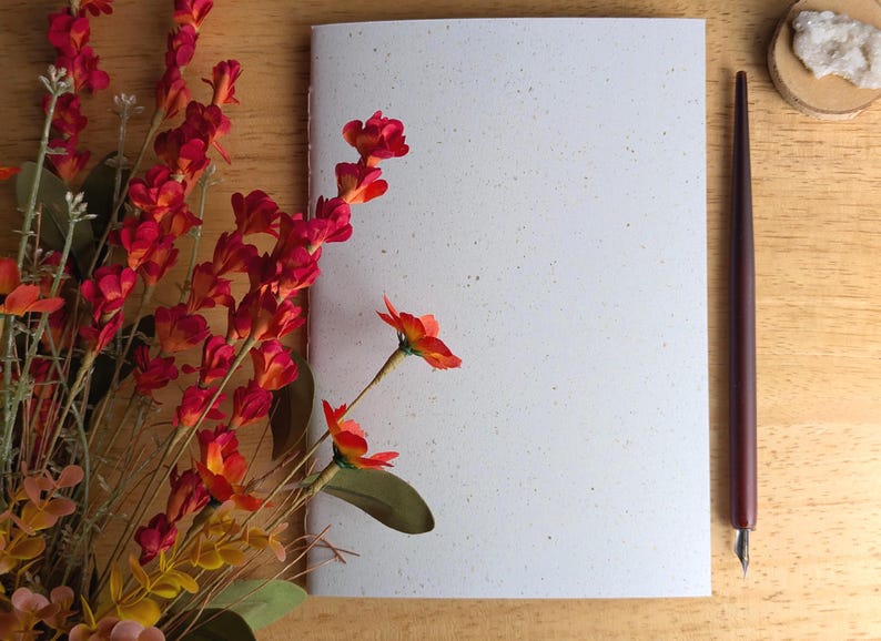 May include: A white notebook with a speckled texture, positioned next to a bouquet of vibrant red and orange flowers and a dark brown calligraphy pen with a metal nib. The notebook features a clean, blank cover and a stitched spine detail.
