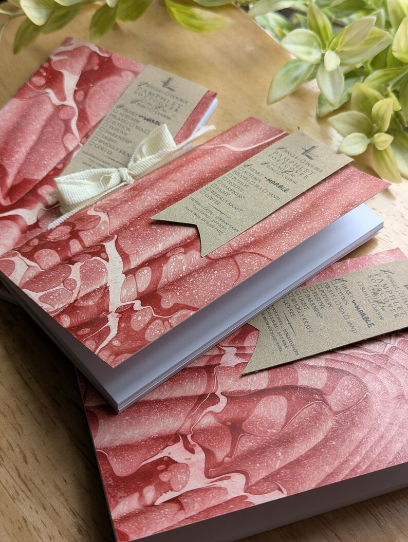 May include: Notebooks with red and white marbled covers. Each notebook has a kraft paper tag attached with a white ribbon. The tags list options such as "Autumn", "Darkness", and "Coffee".