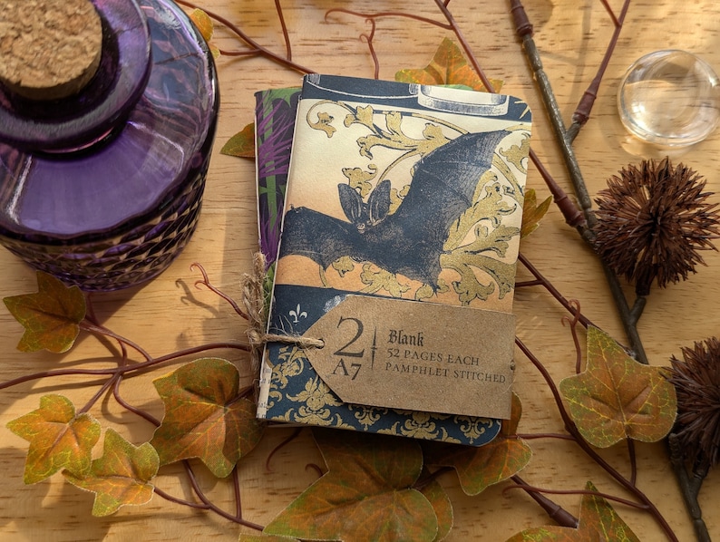 May include: Two A7 blank notebooks, pamphlet stitched, tied with twine. The covers feature a bat illustration with gold accents and a dark blue floral pattern. A purple glass bottle and decorative foliage are in the background.