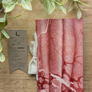 May include: A softcover pamphlet with a marbled red and white design, tied with a cream ribbon. A small tag with text is visible. The pamphlet is made from recycled paper.