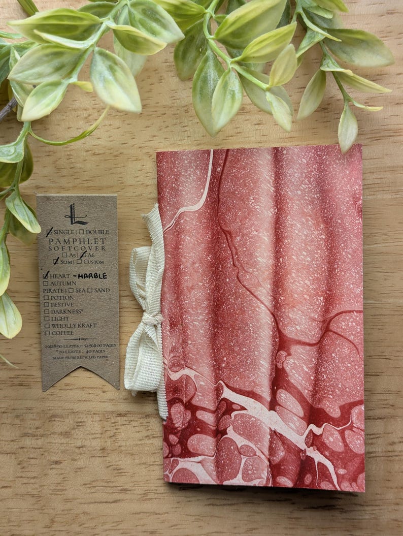 May include: A softcover pamphlet with a marbled red and white design, tied with a cream ribbon. A small tag with text is visible. The pamphlet is made from recycled paper.