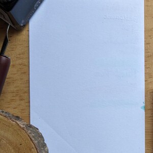 May include: A blank, light blue rectangular card with the embossed word "Squiggles" in the upper right corner. A black ink bottle and a pen are visible in the upper left corner. The card is on a wooden surface.