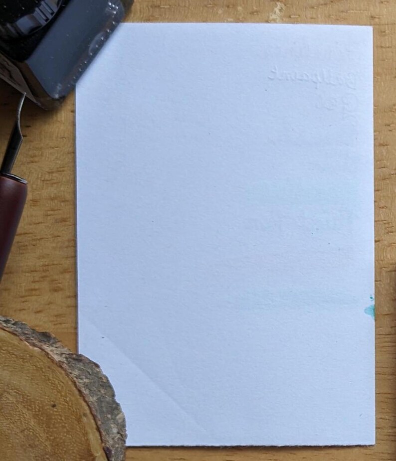 May include: A blank, light blue rectangular card with the embossed word "Squiggles" in the upper right corner. A black ink bottle and a pen are visible in the upper left corner. The card is on a wooden surface.