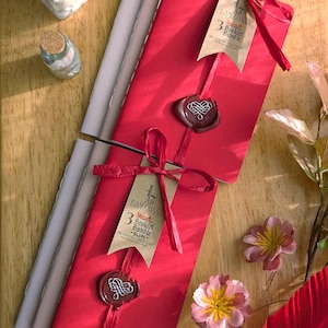 May include: A red booklet bundle tied with a ribbon and sealed with wax. The tag reads "3 Heart Booklet Bundle - SLIM". The image also shows small jars and decorative flowers.