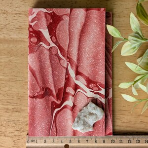 May include: A red and white marbled notebook with a small, light-colored crystal cluster resting on top. A ruler shows measurements in inches. The notebook's design features flowing patterns, and the crystal adds a natural element.