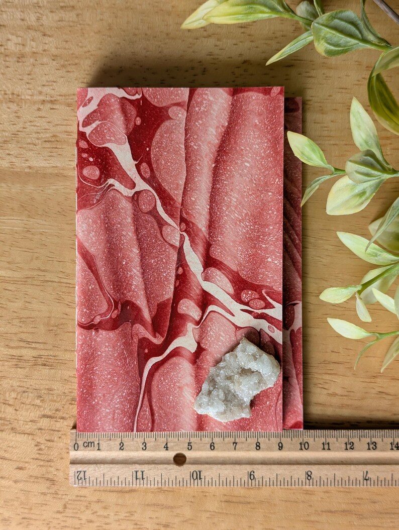 May include: A red and white marbled notebook with a small, light-colored crystal cluster resting on top. A ruler shows measurements in inches. The notebook's design features flowing patterns, and the crystal adds a natural element.