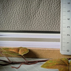 May include: A stack of layered materials including a top sheet of textured light grey leather, followed by thin layers of black, white, and beige paper, resting on a brown board. A white ruler shows a thickness of approximately 2.5 centimetres.
