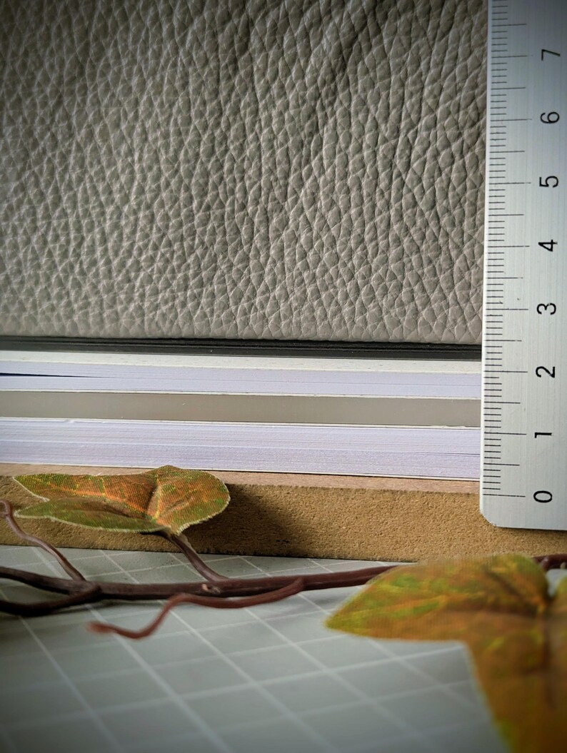 May include: A stack of layered materials including a top sheet of textured light grey leather, followed by thin layers of black, white, and beige paper, resting on a brown board. A white ruler shows a thickness of approximately 2.5 centimetres.