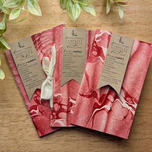 May include: Three red and white marbled pamphlet softcovers with brown tags and a cream ribbon on a wooden surface. The tags have text and a logo. Green leaves are in the upper left corner.
