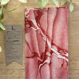May include: A softcover pamphlet with a red and white marbled design. The pamphlet has a vertical orientation and is next to a small tag with text. The tag lists options such as "Heart Marble" and "Autumn". The pamphlet is made from recycled paper.