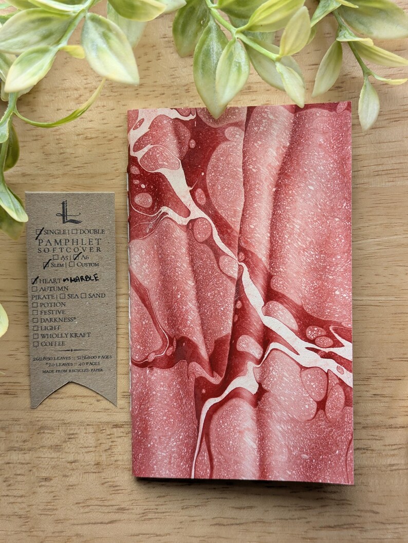 May include: A softcover pamphlet with a red and white marbled design. The pamphlet has a vertical orientation and is next to a small tag with text. The tag lists options such as "Heart Marble" and "Autumn". The pamphlet is made from recycled paper.