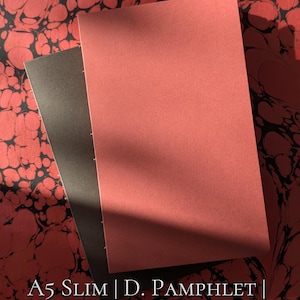 May include: Two slim pamphlets with solid covers in muted red and dark grey, featuring visible side stitching. The text reads: A5 SLIM | D. PAMPHLET | 20.6CM X 13 X .6CM ::PR[XL] & LC[M] COMPATIBLE REFILL::. The items are set against a red and black marbled pattern.