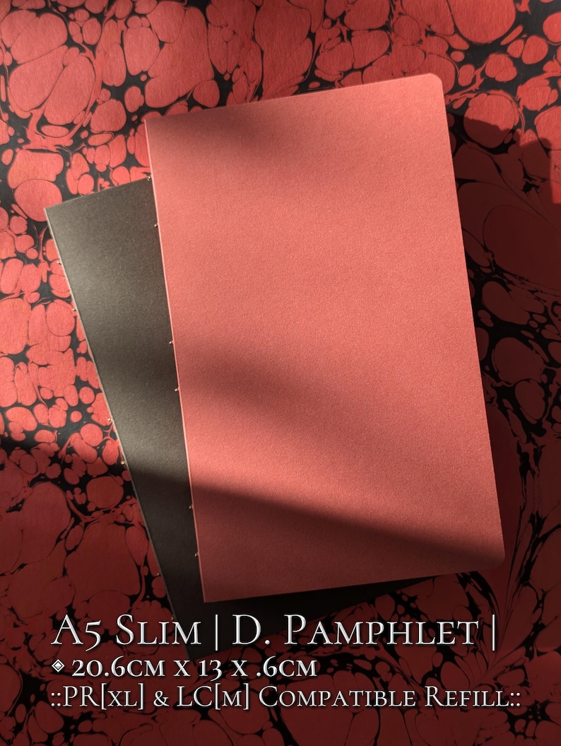 May include: Two slim pamphlets with solid covers in muted red and dark grey, featuring visible side stitching. The text reads: A5 SLIM | D. PAMPHLET | 20.6CM X 13 X .6CM ::PR[XL] & LC[M] COMPATIBLE REFILL::. The items are set against a red and black marbled pattern.