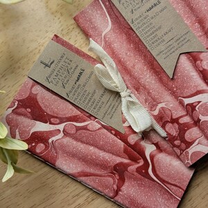 May include: Two softcover pamphlets with a red and white marbled design, tied with a cream ribbon. Each pamphlet has a brown tag with text, including "Heart Marble" and a list of options. The pamphlets are on a wooden surface.