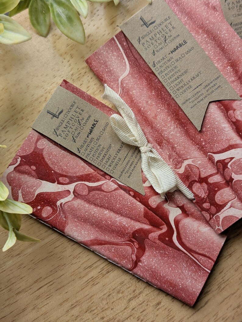 May include: Two softcover pamphlets with a red and white marbled design, tied with a cream ribbon. Each pamphlet has a brown tag with text, including "Heart Marble" and a list of options. The pamphlets are on a wooden surface.