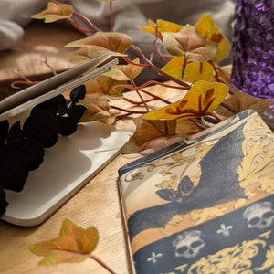 May include: Two open notebooks with decorative covers. One features a black butterfly design, the other a blue and gold pattern with skulls and fleur-de-lis. Autumn leaves and a purple glass vase are in the background, creating a seasonal aesthetic.