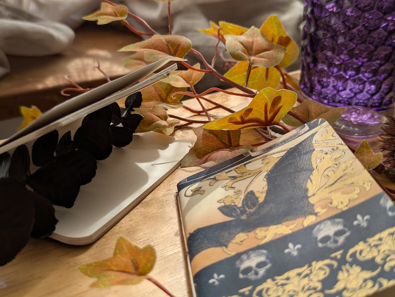 May include: Two open notebooks with decorative covers. One features a black butterfly design, the other a blue and gold pattern with skulls and fleur-de-lis. Autumn leaves and a purple glass vase are in the background, creating a seasonal aesthetic.