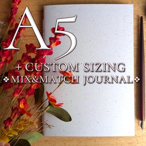 May include: A white A5 journal with a speckled texture, featuring the text A5 + CUSTOM SIZING MIX&MATCH JOURNAL. The journal is positioned next to a dark brown calligraphy pen and a cluster of red and orange artificial flowers on a wooden surface.