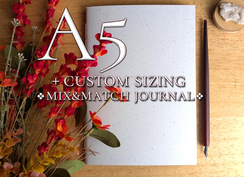 May include: A white A5 journal with a speckled texture, featuring the text A5 + CUSTOM SIZING MIX&MATCH JOURNAL. The journal is positioned next to a dark brown calligraphy pen and a cluster of red and orange artificial flowers on a wooden surface.