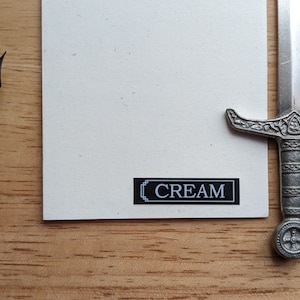 May include: A cream-colored card with the word "CREAM" in a black and white rectangular box. A silver letter opener shaped like a sword and a brown pen are also in the image.