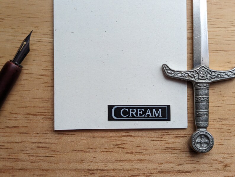 May include: A cream-colored card with the word "CREAM" in a black and white rectangular box. A silver letter opener shaped like a sword and a brown pen are also in the image.