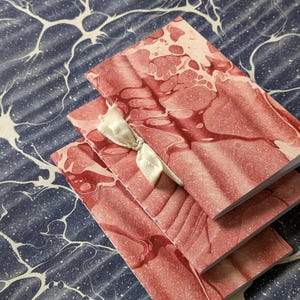 May include: Three small notebooks with red and white marbled covers are stacked on a blue and white patterned surface. A cream-colored ribbon is tied around the notebooks. The notebooks have a soft, organic design.