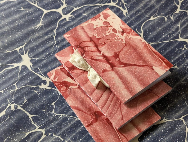 May include: Three small notebooks with red and white marbled covers are stacked on a blue and white patterned surface. A cream-colored ribbon is tied around the notebooks. The notebooks have a soft, organic design.