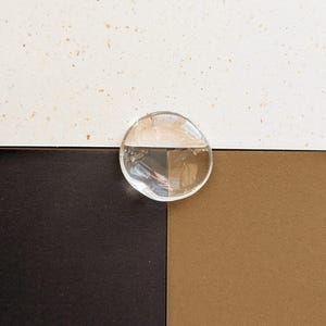 May include: A clear, circular glass disc positioned over a background divided into three sections: a speckled off-white top, a solid dark brown bottom left, and a solid tan bottom right. The text LIGHT ROAST, COCONUT, and HAZELNUT are visible.