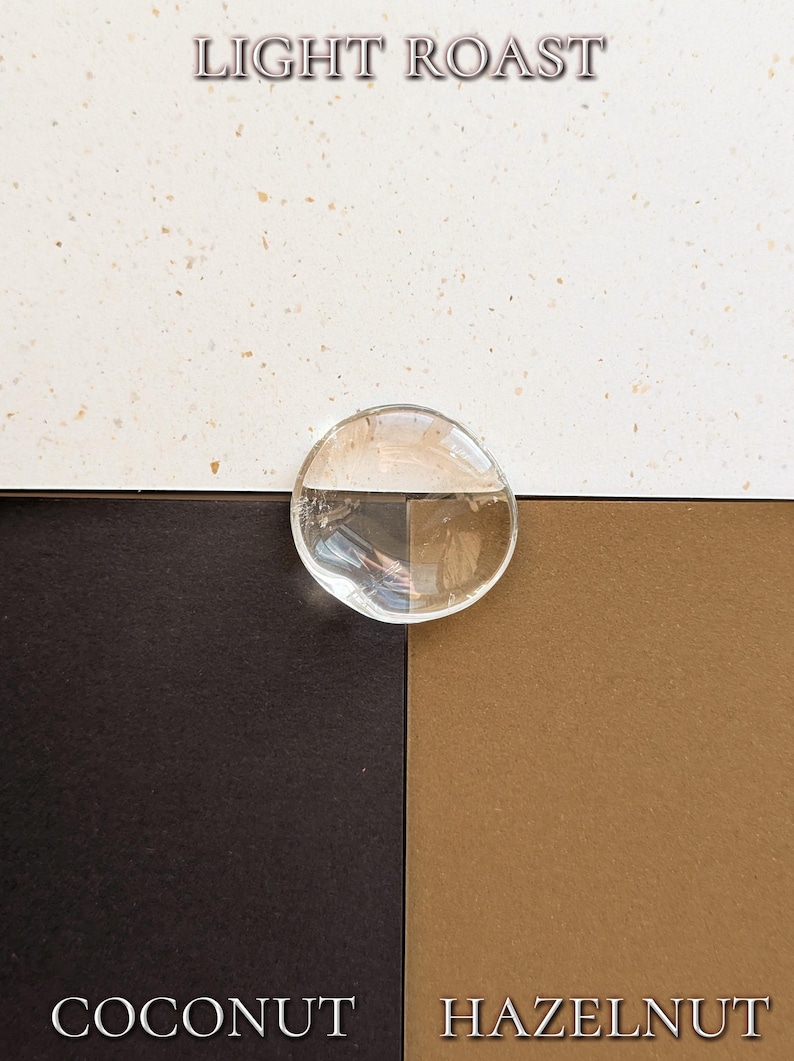 May include: A clear, circular glass disc positioned over a background divided into three sections: a speckled off-white top, a solid dark brown bottom left, and a solid tan bottom right. The text LIGHT ROAST, COCONUT, and HAZELNUT are visible.