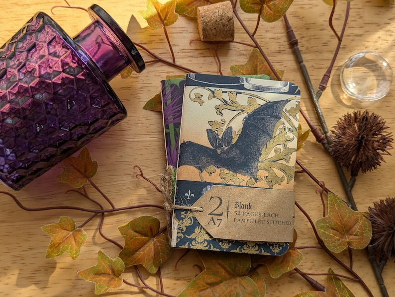 May include: A stack of two A7 blank notebooks with a bat illustration, tied with twine. The notebooks have a vintage aesthetic with a dark blue and gold color scheme. A purple glass bottle and autumn leaves are in the background.