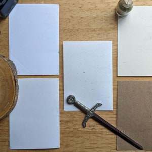 May include: An overhead shot of stationery supplies on a wooden surface. Includes a calligraphy pen, ink bottle, small glass bottle, and a decorative sword letter opener. There are also several blank white and brown rectangular cards.