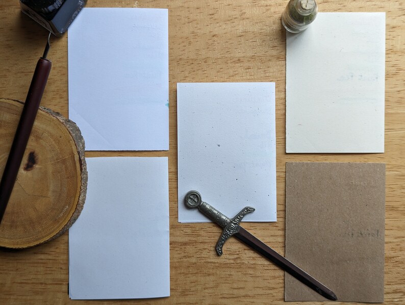 May include: An overhead shot of stationery supplies on a wooden surface. Includes a calligraphy pen, ink bottle, small glass bottle, and a decorative sword letter opener. There are also several blank white and brown rectangular cards.