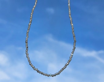 May include: A silver necklace made of small, round beads. The necklace is hanging against a blue sky with white clouds.