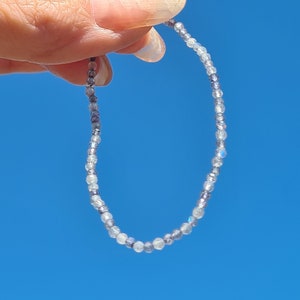 Könnte beinhalten: Ein zartes Armband aus kleinen, irisierenden, hellblauen und grauen Perlen. Das Armband ist auf einer durchsichtigen Gummikordel aufgefädelt und wird vor einem strahlend blauen Himmel gezeigt.