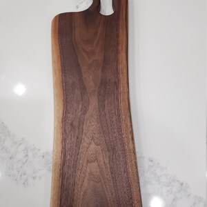 Puede incluir: Una tabla de cortar de madera marrón oscuro grande con un mango asimétrico único. La tabla está hecha de madera de nogal y tiene un acabado natural de borde vivo.