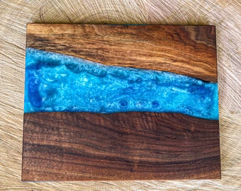 May include: A wooden cutting board with a blue epoxy resin inlay. The resin has a wavy, river-like shape and is filled with white and blue glitter.
