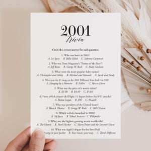 May include: A white card with the title "2001 Trivia" in a decorative font. The card features trivia questions about the year 2001, including pop culture, news, and technology. The card is designed for a game or party.
