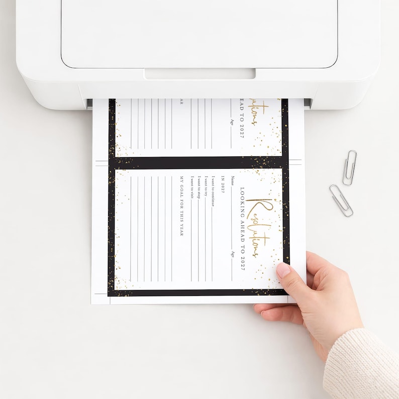 May include: A white printer outputting a sheet of paper with two identical black and gold goal-setting templates. The text reads Resolutions Looking Ahead To 2027, with sections for name, age, and goals. Two silver metal paper clips lie nearby.
