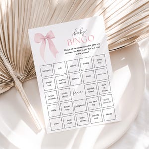 May include: A pink and white baby shower bingo card with a pink bow at the top. The card has a grid of squares with different baby-related items listed in each square, such as a crib, onesie, diaper, and rattle.
