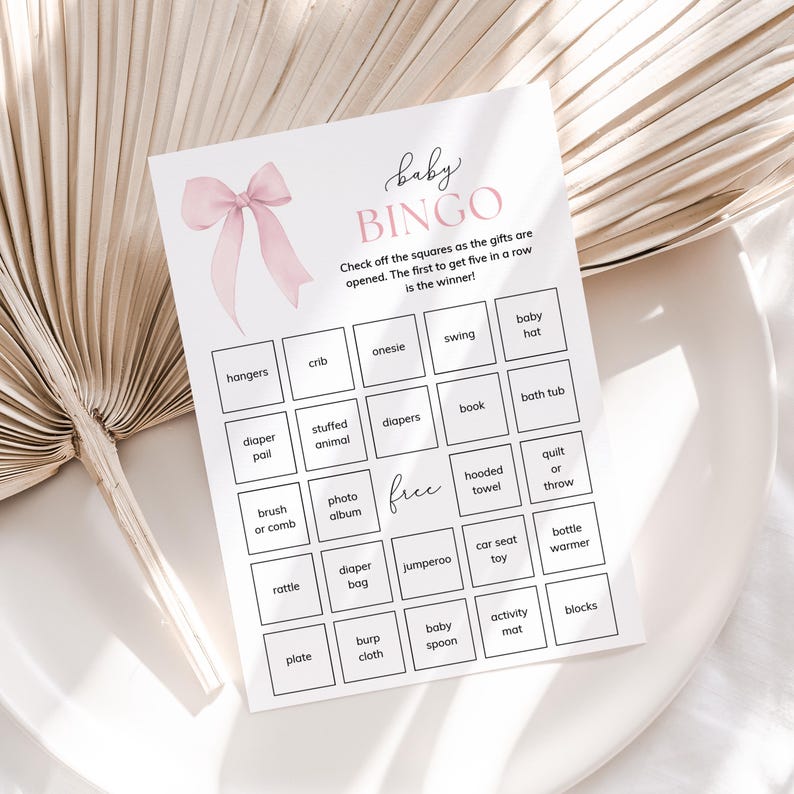 May include: A pink and white baby shower bingo card with a pink bow at the top. The card has a grid of squares with different baby-related items listed in each square, such as a crib, onesie, diaper, and rattle.