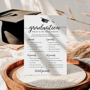 May include: A graduation party game card with the text "graduation WHAT'S ON YOUR PHONE". The card lists point values for various phone-related activities. A graduation cap is at the top. The card is on a wooden tray.