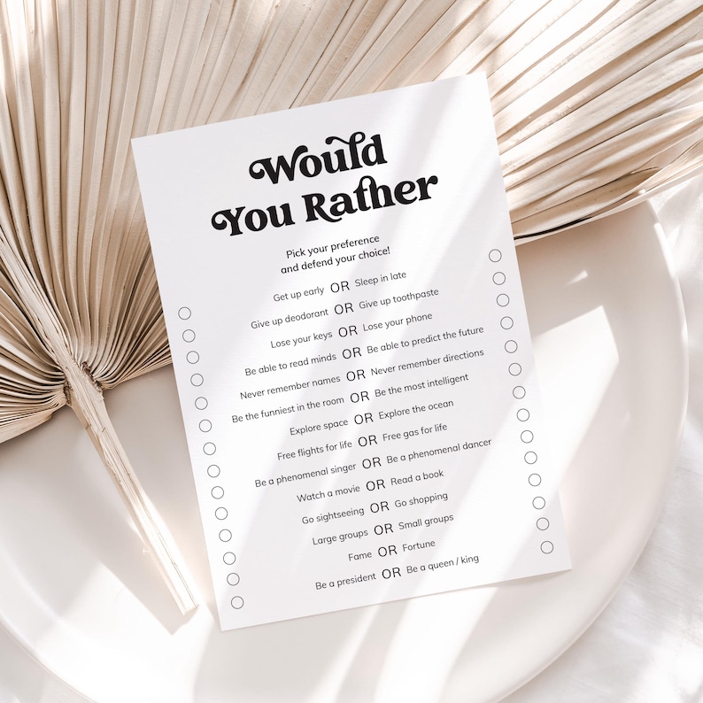 May include: A white card with the text "Would You Rather" in a bold, retro font. The card lists various "Would you rather" questions with check boxes. The card is on a white plate with a dried palm leaf in the background.