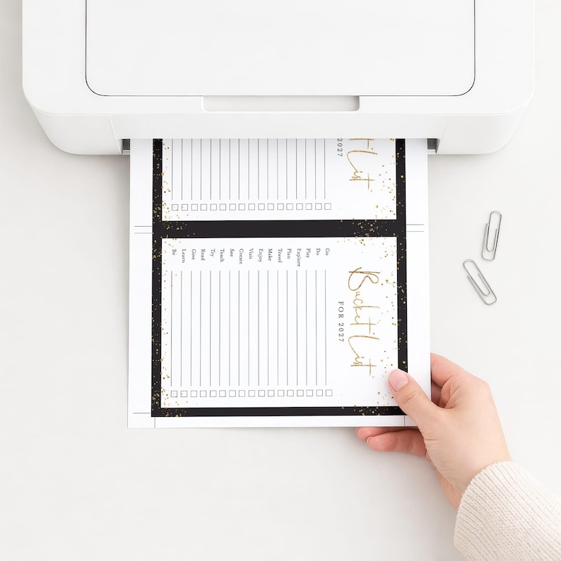 K&ouml;nnte beinhalten: Ein wei&szlig;er Drucker, der ein Blatt Papier mit zwei identischen 2027 Bucket-List-Vorlagen ausgibt. Jede Vorlage hat einen schwarzen Rand mit goldenen Sprenkeln, einen goldenen Schriftzug Bucket List FOR 2027, eine Liste von Verben und K&auml;stchen.