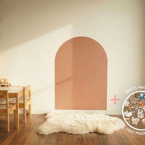 Ein Kinderspielzimmer mit Holzmöbeln, darunter ein Tisch und Stühle, und ein Regal mit Spielzeug. Ein großes, bogenförmiges Wandelement ist in einem gedämpften Korallenfarbton gestrichen. Ein weißer Schaffellteppich liegt auf dem Holzboden.